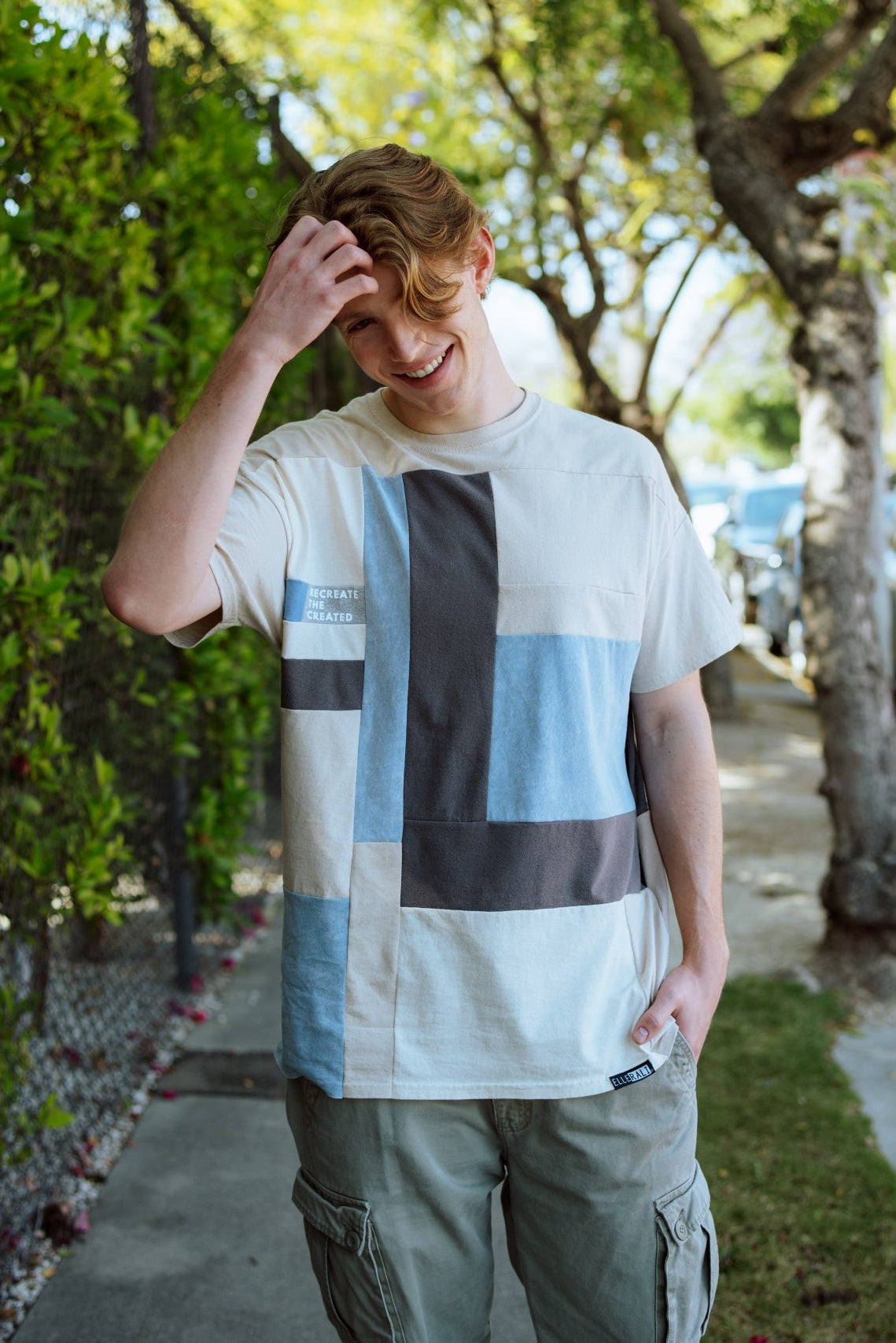 A young man standing on a tree-lined sidewalk wearing a white t-shirt with a large geometric block pattern in light blue, dark gray, and beige, paired with olive cargo pants.