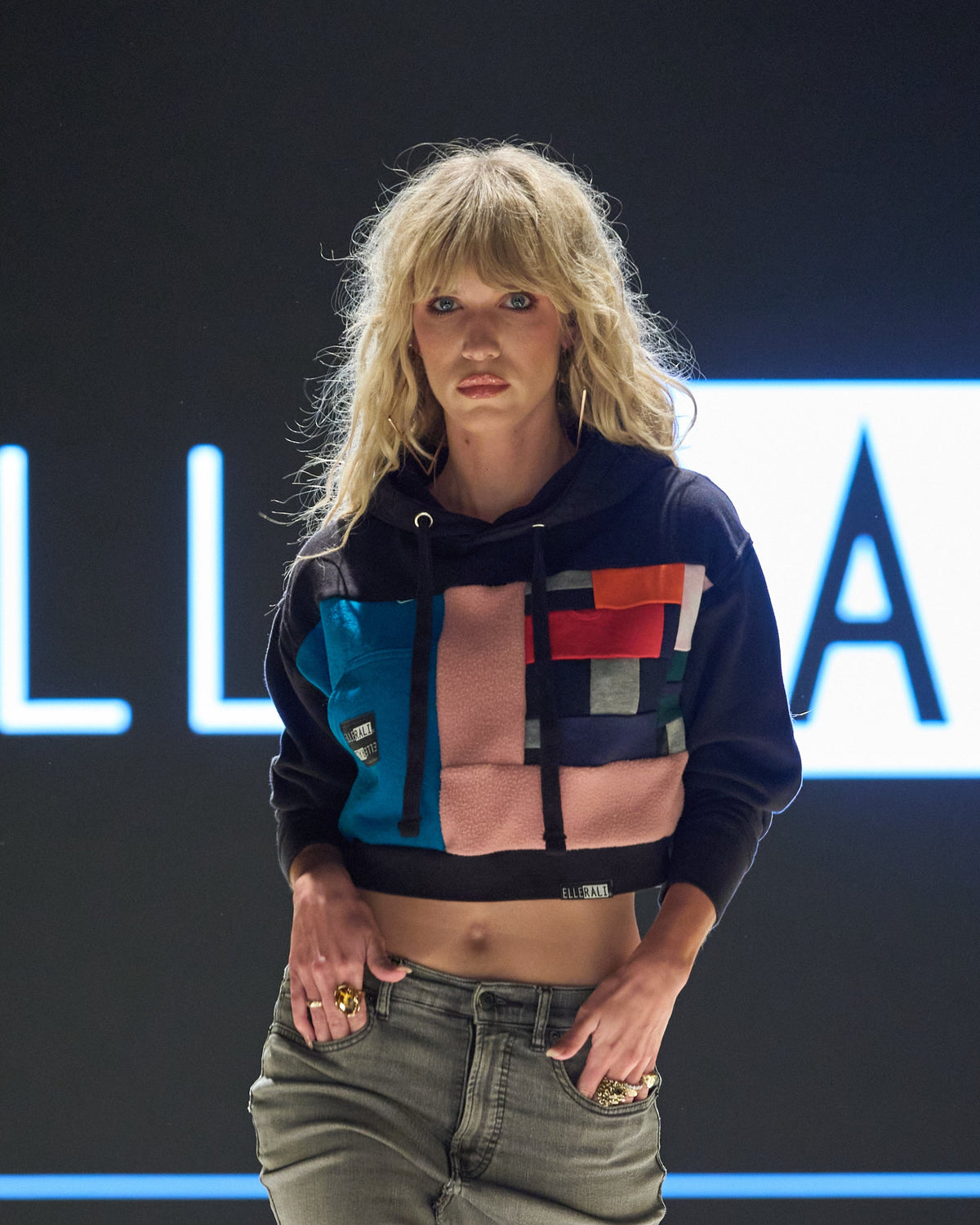 A model walks a fashion runway wearing a cropped multicolor hoodie, a high-waisted olive-gray asymmetrical skirt, and black ankle boots. The background features a large screen with the brand name 'ELLE VALI' in glowing white letters.